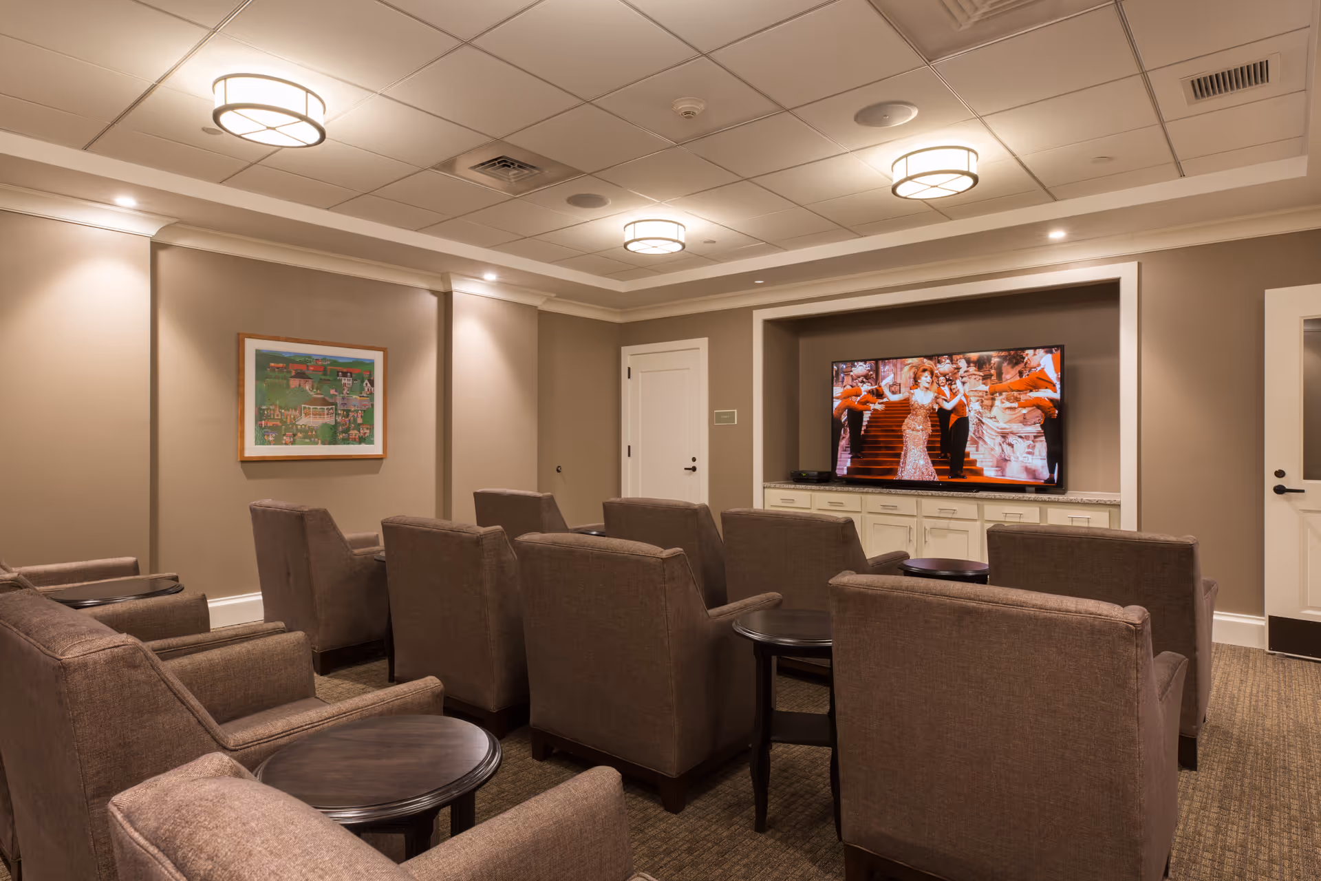 A cozy media room with several brown upholstered armchairs arranged in rows facing a large flat-screen TV mounted on a beige wall. The TV is displaying a scene with people in colorful costumes. The room has beige walls, a carpeted floor, recessed lighting, and two round ceiling lights. There is a framed picture on the left wall and two white doors on the right side of the room.