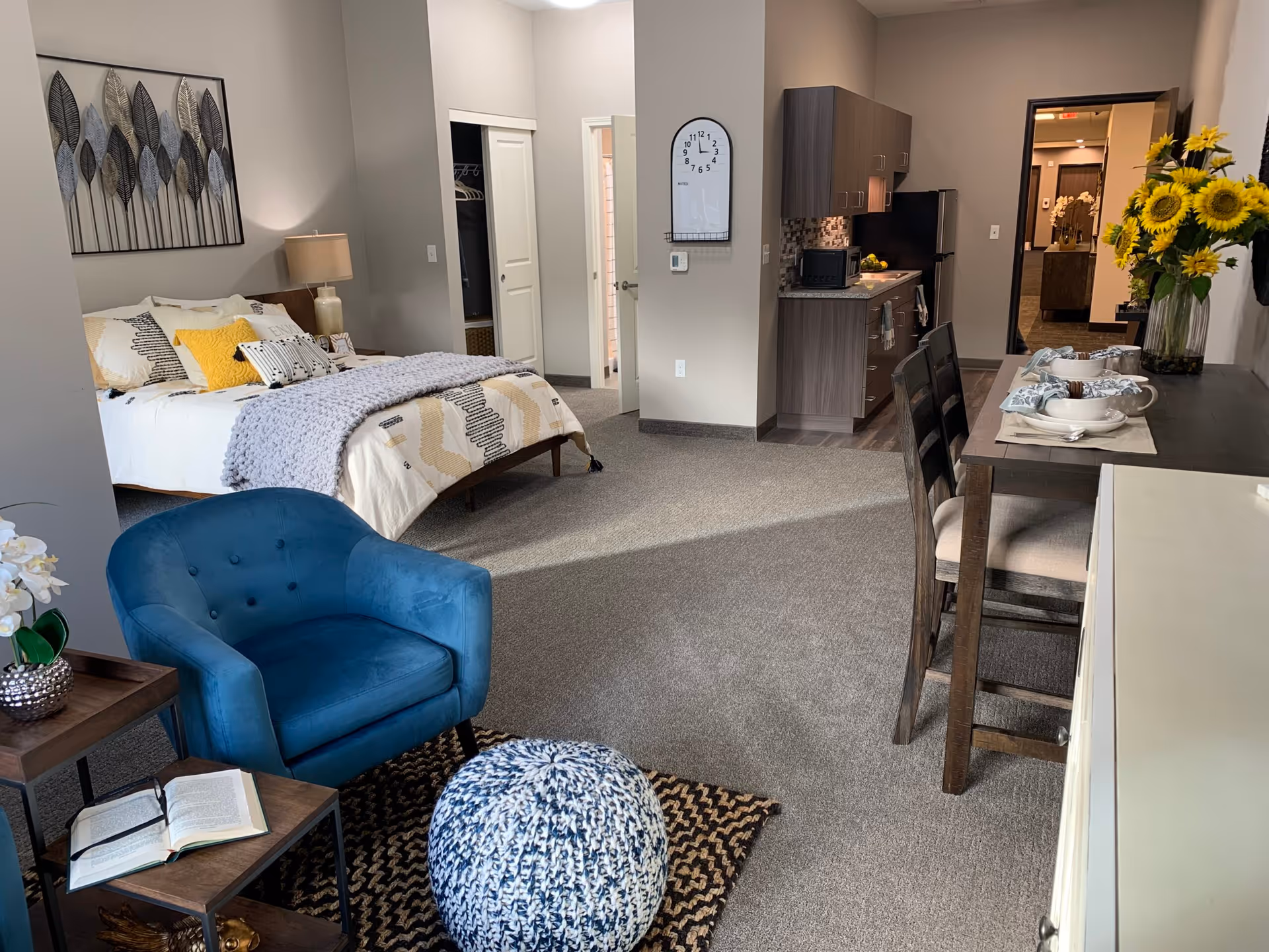 Furnished studio apartment interior showing a bed, blue armchair, dining table, and small kitchenette.