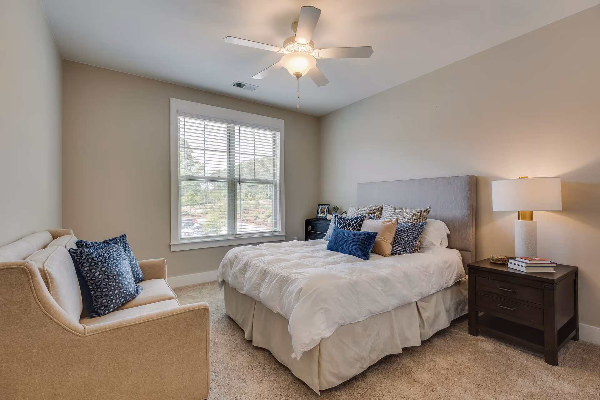 A cozy bedroom with a large bed featuring multiple pillows, a beige sofa with blue patterned cushions, a window with white blinds, a ceiling fan with light, and a dark wooden nightstand with a lamp and books.