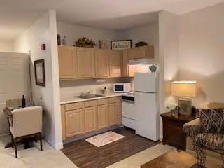 A small kitchen area in a senior living facility featuring light wood cabinets, a white refrigerator, a microwave, a stove, and a sink. To the right of the kitchen is a wooden side table with a lamp and a patterned armchair. To the left, there is a desk with a chair and some decorative items on the wall.