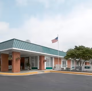 Exterior view of Monroe Health and Rehabilitation Center showing a single-story brick building with a green metal roof, an American flag on a flagpole in front, and a paved driveway with a covered entrance.