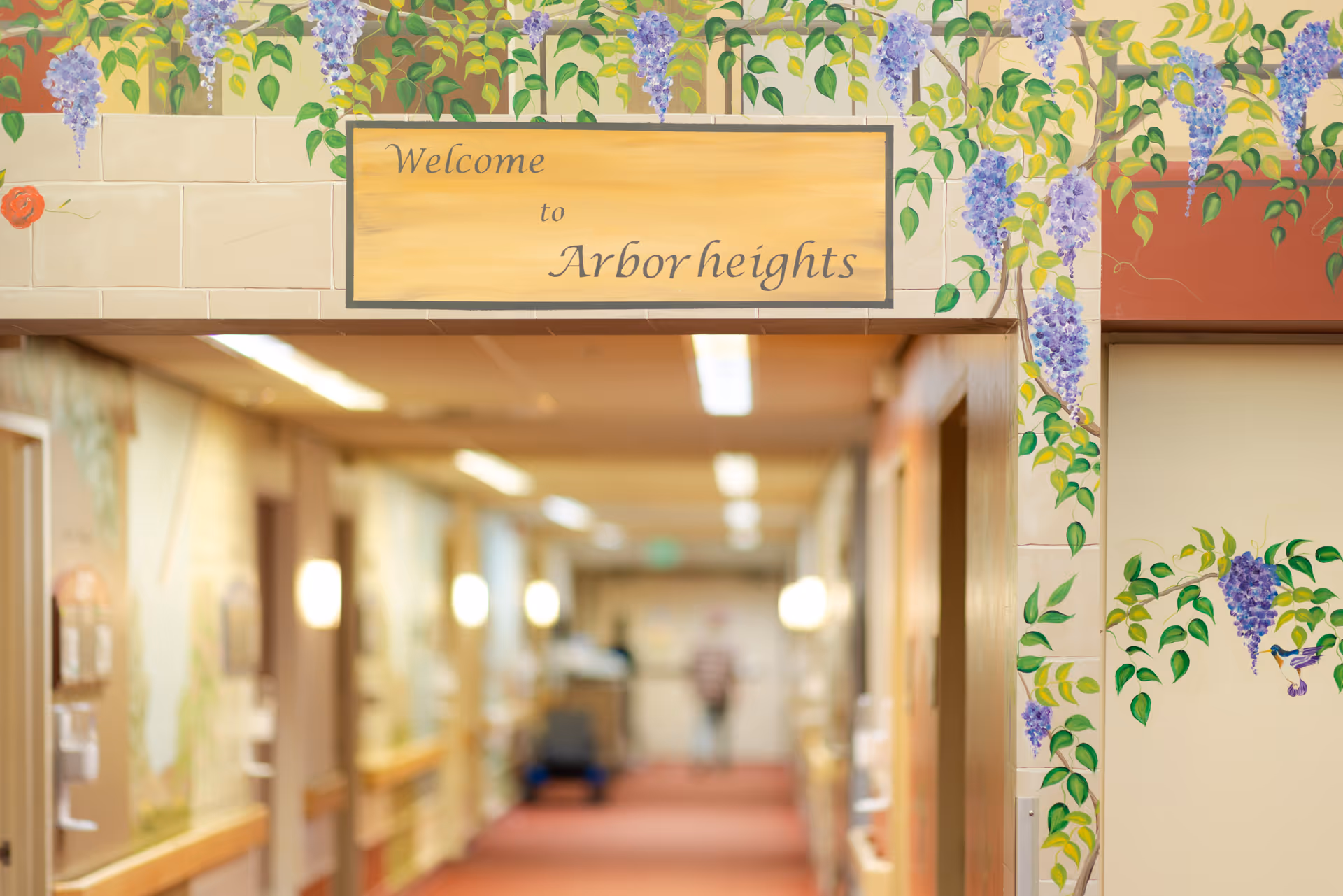 A hallway in a senior living facility with a sign above the entrance that reads 'Welcome to Arbor heights'. The walls around the entrance are decorated with painted purple wisteria flowers and green leaves. The hallway is softly lit with wall sconces and ceiling lights, and there is a person blurred in the distance.