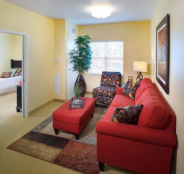 A cozy living room with a red sofa, matching red ottoman, and a patterned armchair. There is a large window with blinds, a tall green plant in a vase, a table lamp, and a framed artwork on the wall. A doorway leads to a bedroom with a bed and pillows visible.