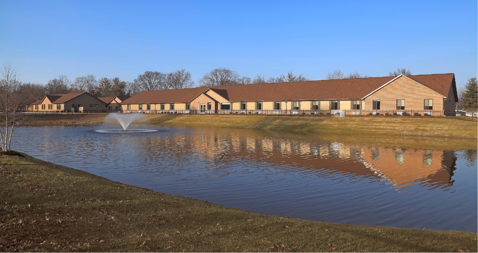 A long, single-story building with a brown roof and beige walls is reflected in a large pond with a water fountain in the center. The scene is outdoors with a clear blue sky and some leafless trees in the background.