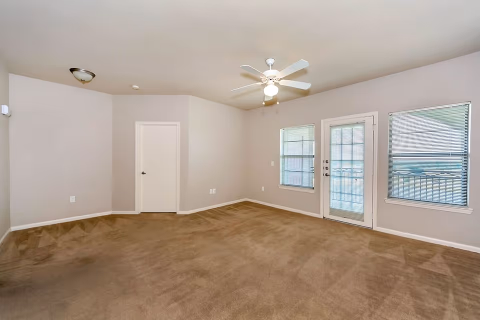 Empty room with beige carpet, light gray walls, a white ceiling fan with light, two windows with blinds, and a glass door leading outside. There is a closed white door on one wall and a ceiling light fixture near the door.