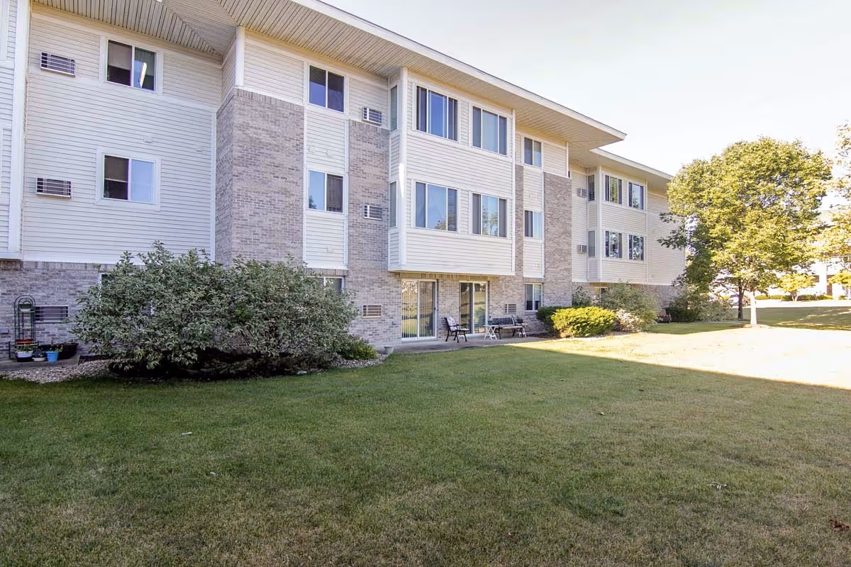 Exterior view of a three-story senior apartment building with a grassy lawn, shrubs, and patio seating.