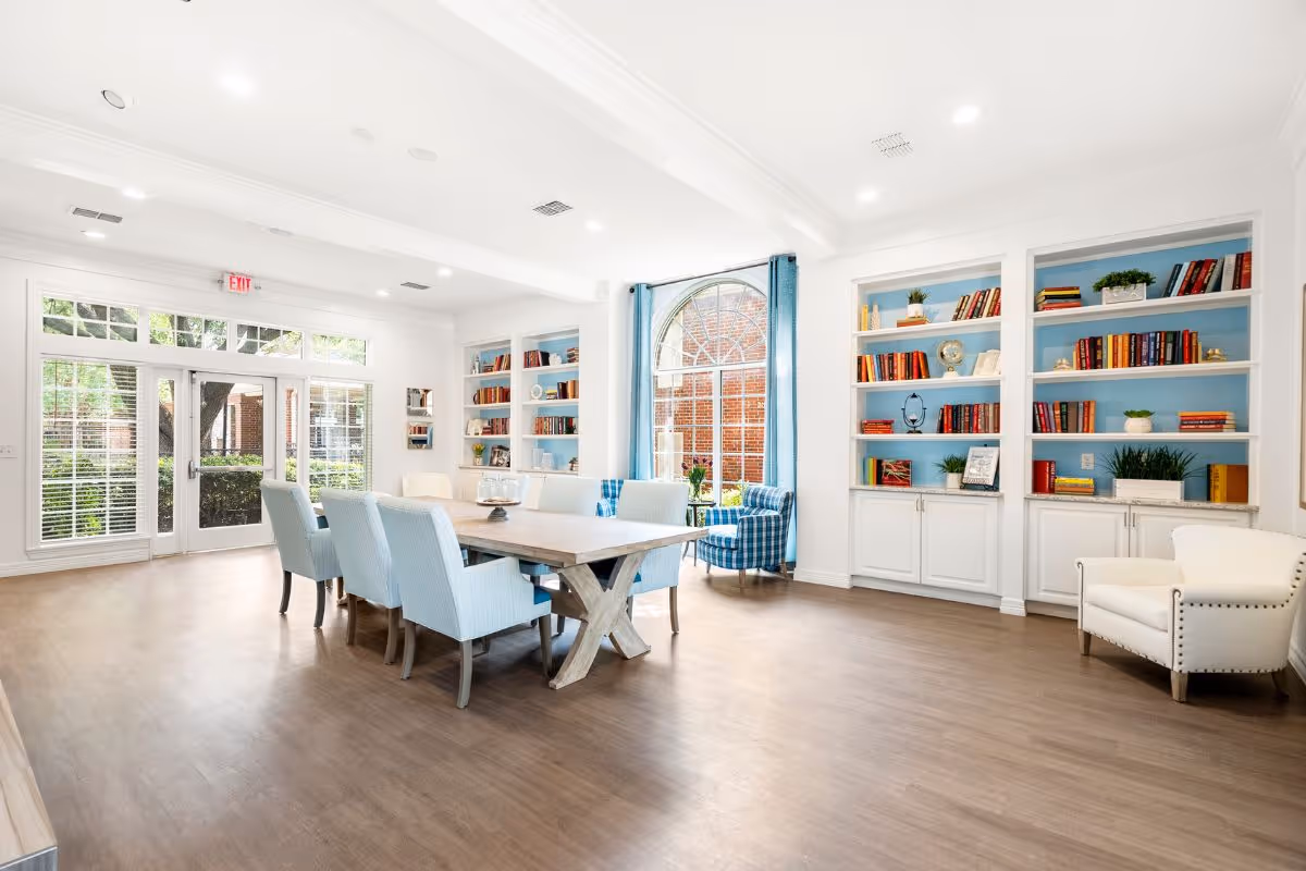 A bright and spacious common area with a large wooden table surrounded by light blue upholstered chairs. The room features built-in white bookshelves filled with books and decorative items, a large arched window with blue curtains, and additional seating including a white armchair and a blue plaid armchair. Glass doors lead outside to a garden area with greenery visible through the windows.