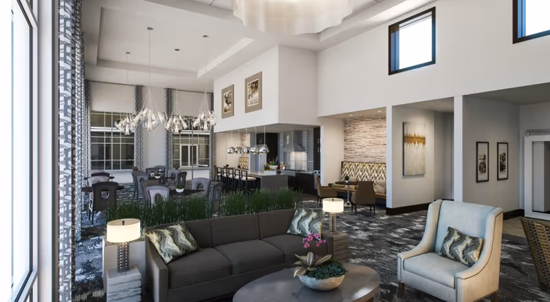 A spacious and modern interior common area featuring a dark gray sofa with patterned pillows, a light-colored armchair, a round coffee table with a plant, and a dining area with multiple tables and chairs. The room has large windows with patterned curtains, contemporary light fixtures, high ceilings with small upper windows, and decorative artwork on the walls.