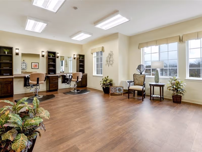 Bright salon-style common room with hair-styling stations, chairs, mirrors, and seating by large windows.