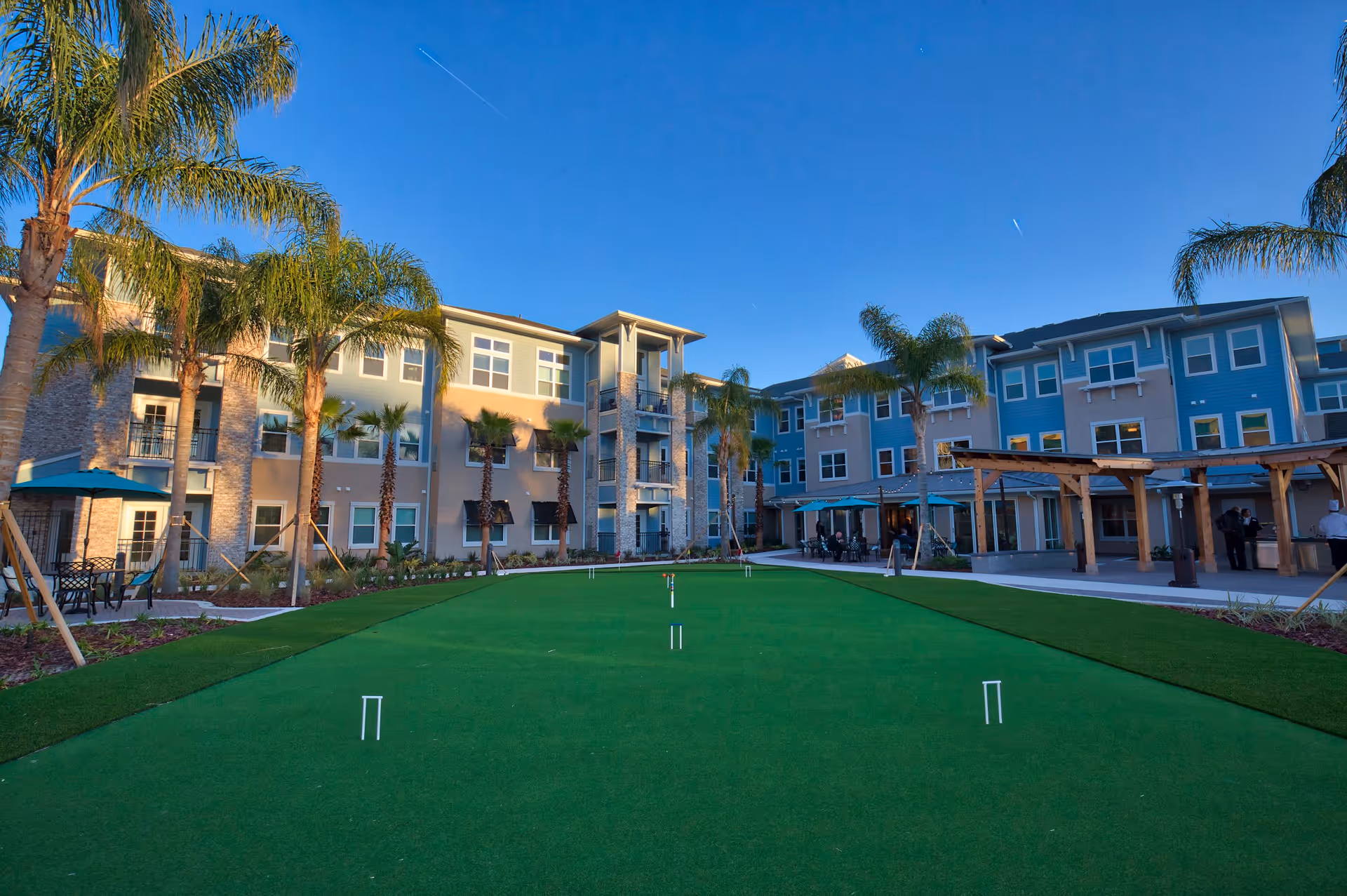 Outdoor courtyard area of a senior living facility with a putting green in the center, surrounded by palm trees and a three-story building with balconies and large windows under a clear blue sky.