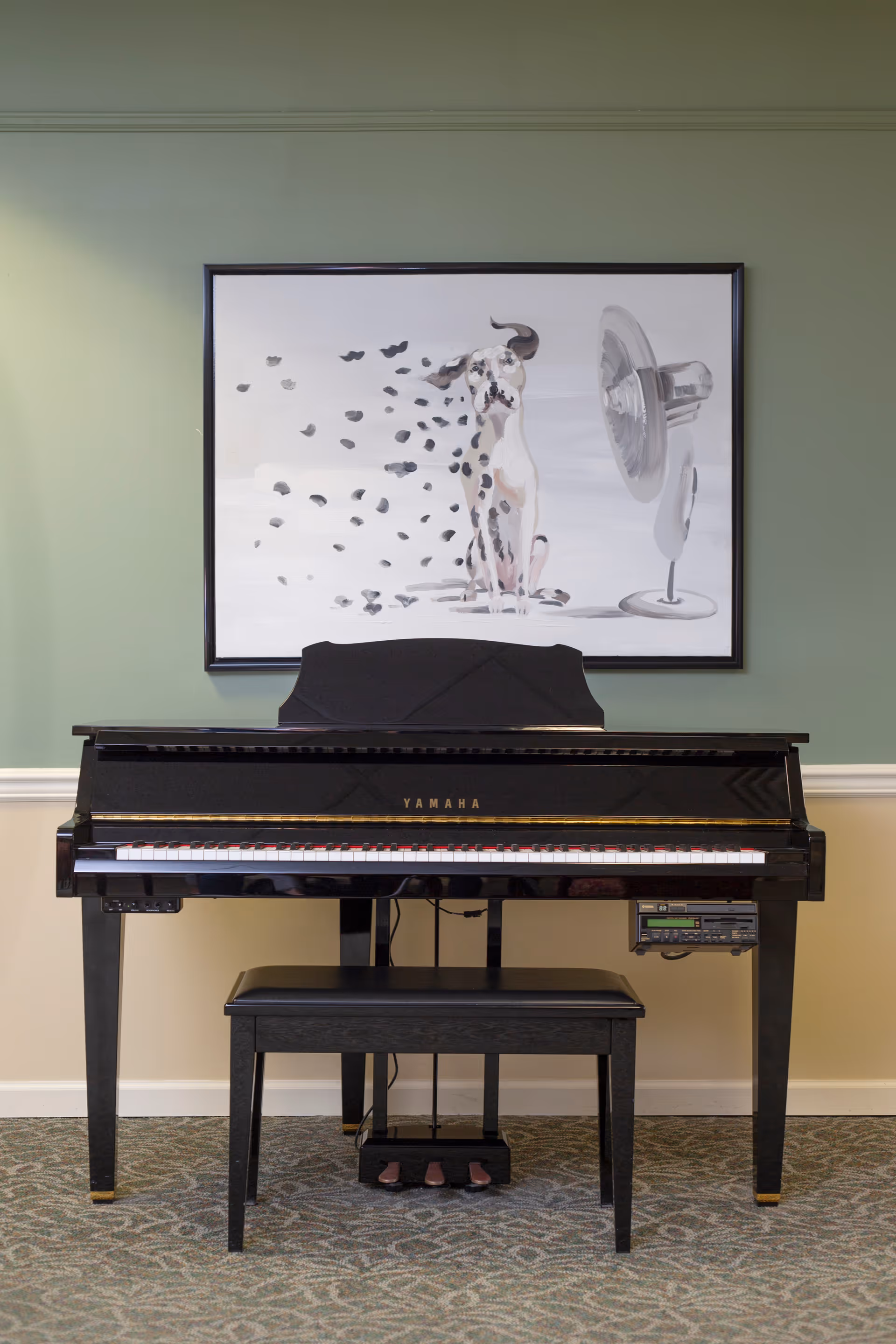 A black Yamaha upright piano with a bench sits against a green wall beneath a framed painting of a dog and a fan.