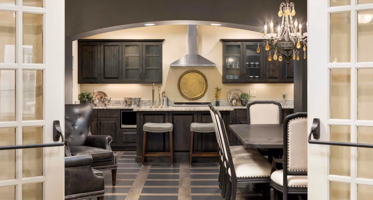 View through open French doors into a kitchen and dining area featuring dark wood cabinetry, granite countertops, a stainless steel range hood, two bar stools at a kitchen island, a dark wood dining table with upholstered chairs, a chandelier with hanging crystals, and two leather armchairs.