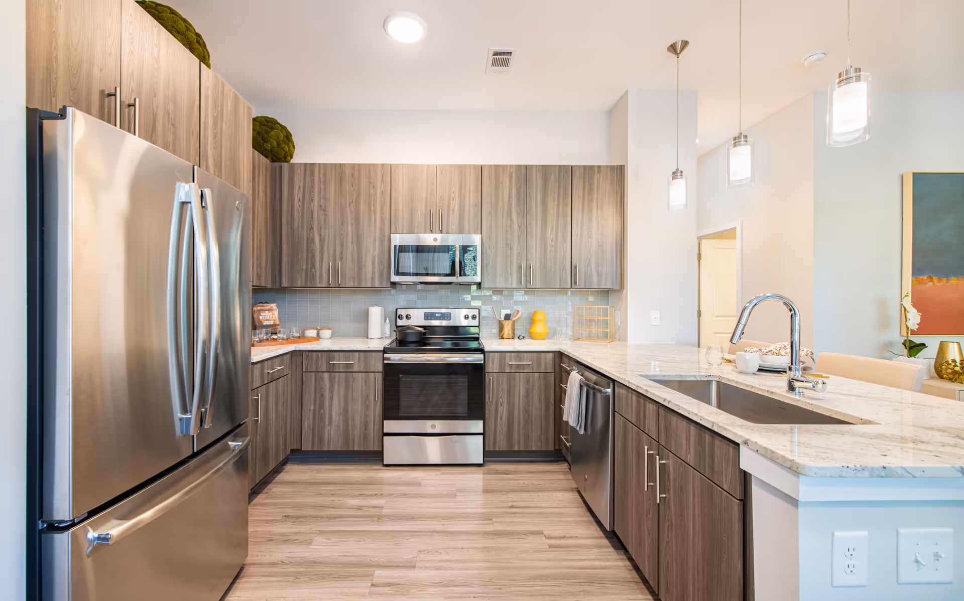 Modern kitchen with stainless steel refrigerator, oven, microwave, and dishwasher. The kitchen features wood cabinets, a marble countertop island with a sink, pendant lighting, and light wood flooring.