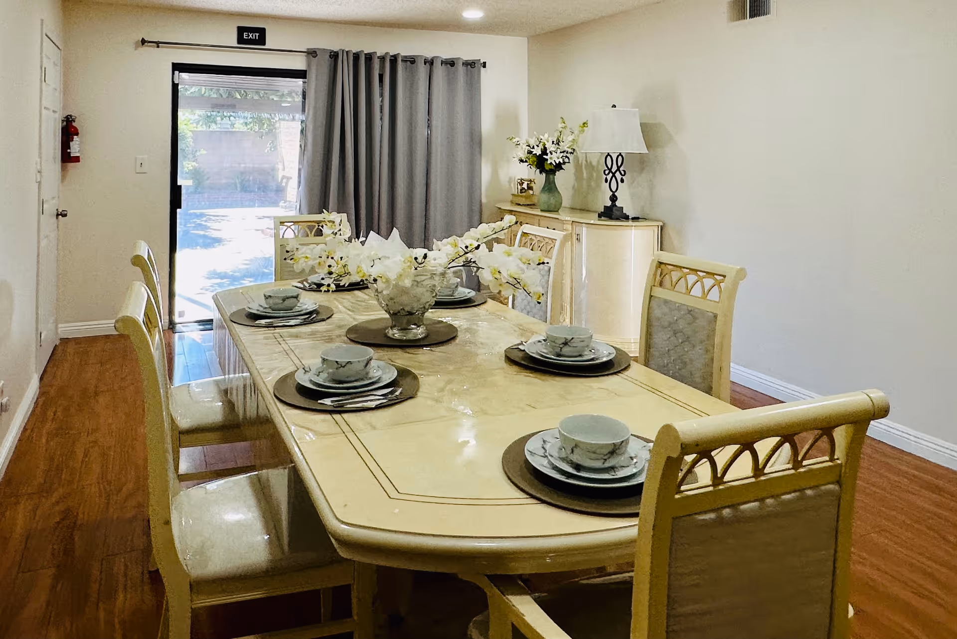A dining room with a rectangular table set for six people. The table has a cream-colored surface with a floral centerpiece and matching dishware including plates, bowls, and cups. Six chairs with cushioned seats surround the table. In the background, there is a sideboard with a lamp and a vase of flowers. A sliding glass door with gray curtains leads to an outdoor area.