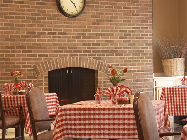 Dining area with tables covered in red and white checkered tablecloths, each table decorated with a small vase of flowers and folded napkins. The room features a brick fireplace with a clock above it and a cabinet with a large basket of dried branches in the background.