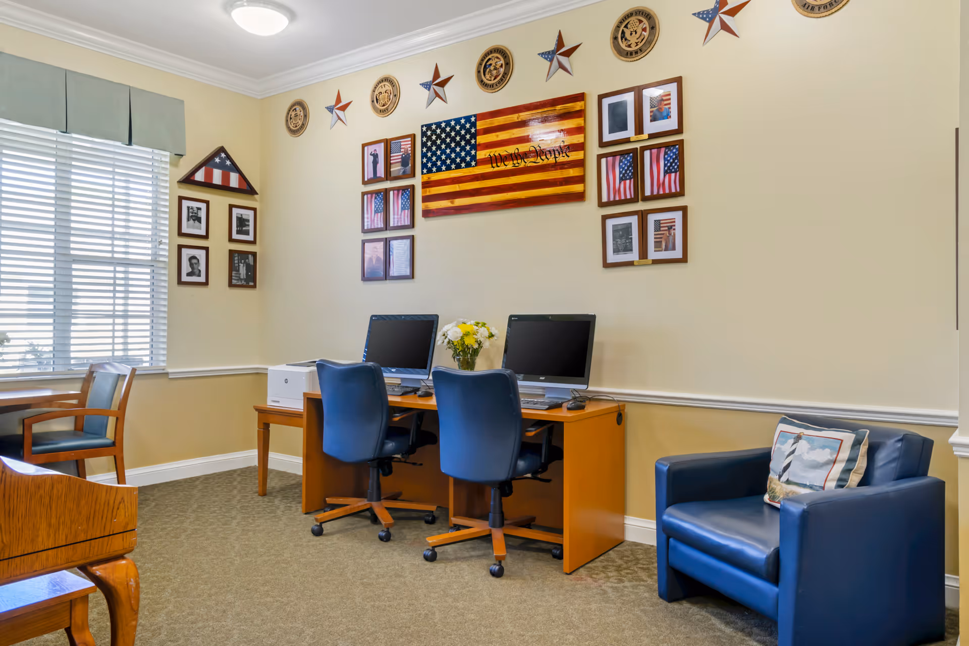 A small computer area in a senior living facility with two desktop computers on a wooden desk, two blue office chairs, a blue armchair with a lighthouse-themed pillow, and patriotic wall decorations including an American flag, stars, and framed photos.