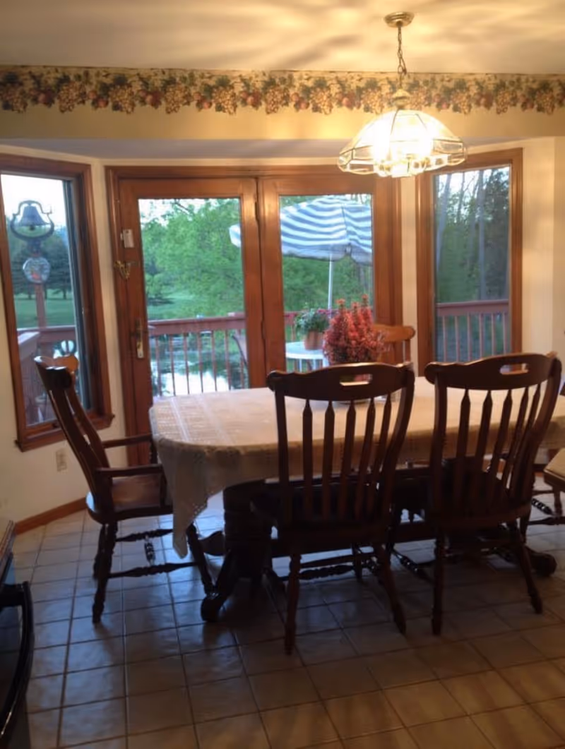A dining area with a wooden table covered by a white tablecloth and surrounded by wooden chairs. The room has large windows and a glass door leading to an outdoor deck with a striped umbrella and greenery visible outside. A hanging light fixture is above the table, and there is a floral wallpaper border near the ceiling.
