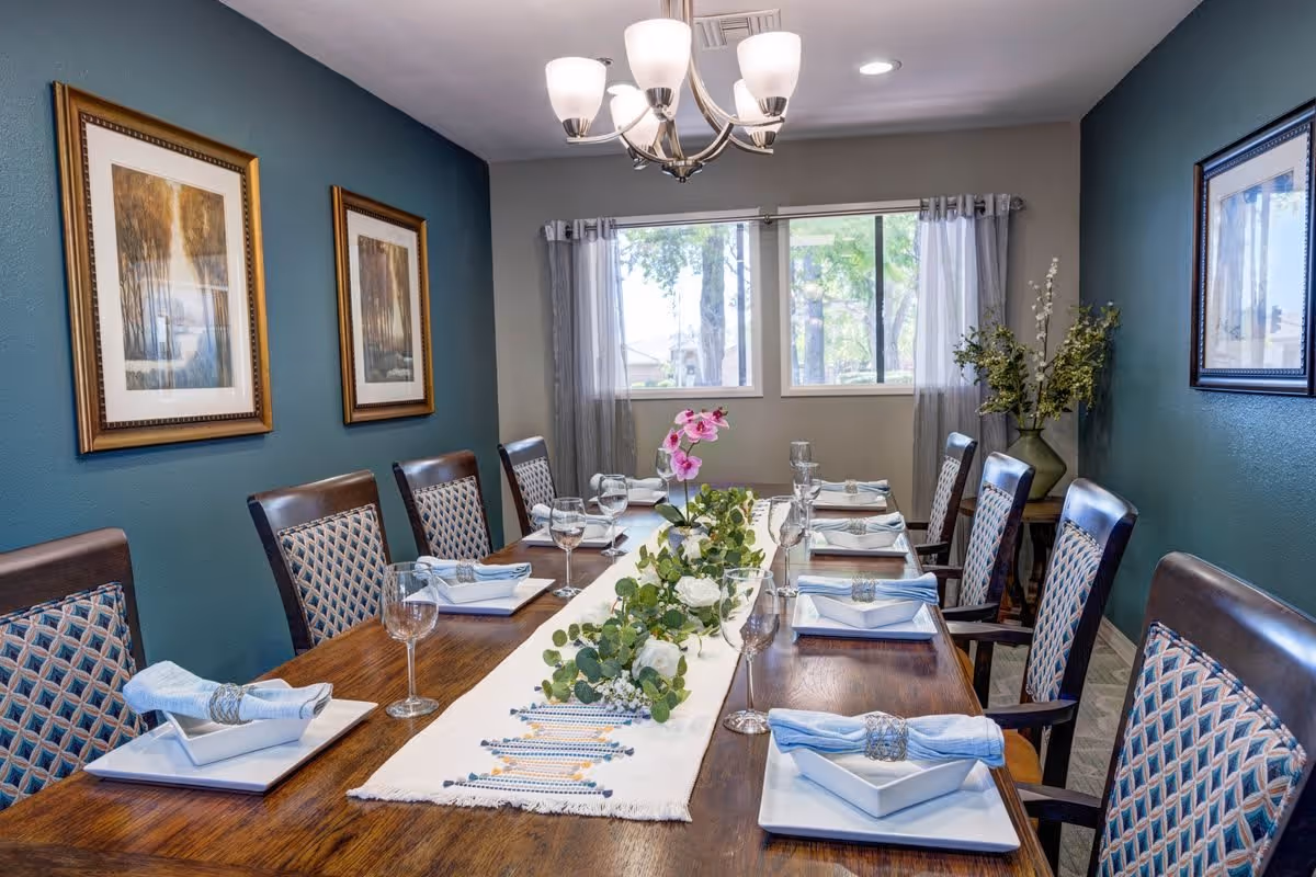 A dining room with a long wooden table set for eight people. Each place setting includes a white square plate, a folded blue napkin with a silver napkin ring, and a wine glass. A white table runner with a floral centerpiece featuring green leaves and white and pink flowers runs down the middle of the table. The room has teal and gray walls, two framed landscape paintings on one wall, a window with sheer curtains, and a chandelier with five lights hanging from the ceiling.