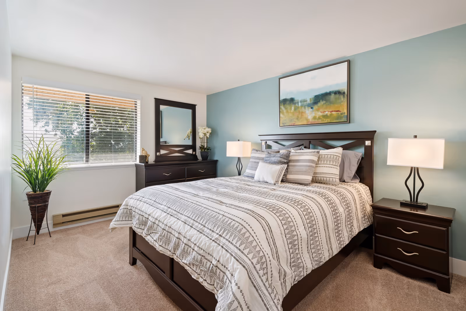 Bright bedroom with a neatly made bed, patterned bedding, two nightstands with lamps, dresser with mirror, and a window.