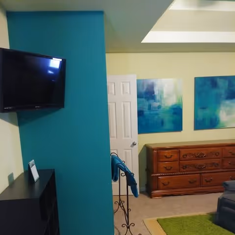 Interior view of a living room area with a wall-mounted flat screen TV on a teal accent wall, a wooden dresser with multiple drawers, two abstract blue and green paintings hanging above the dresser, and a partially visible gray armchair on a green rug.