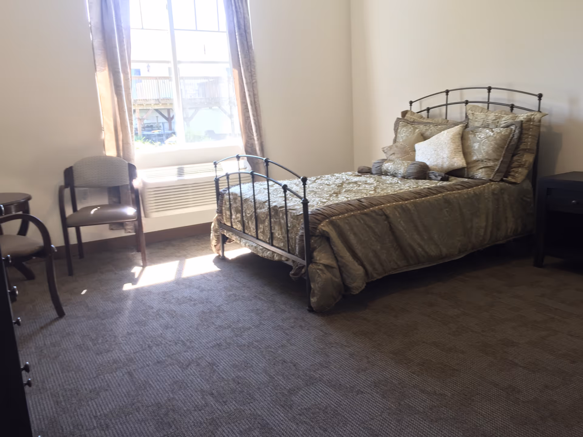 Sunlit bedroom with a single metal-frame bed, decorative pillows, a bedside table and chairs by a window.