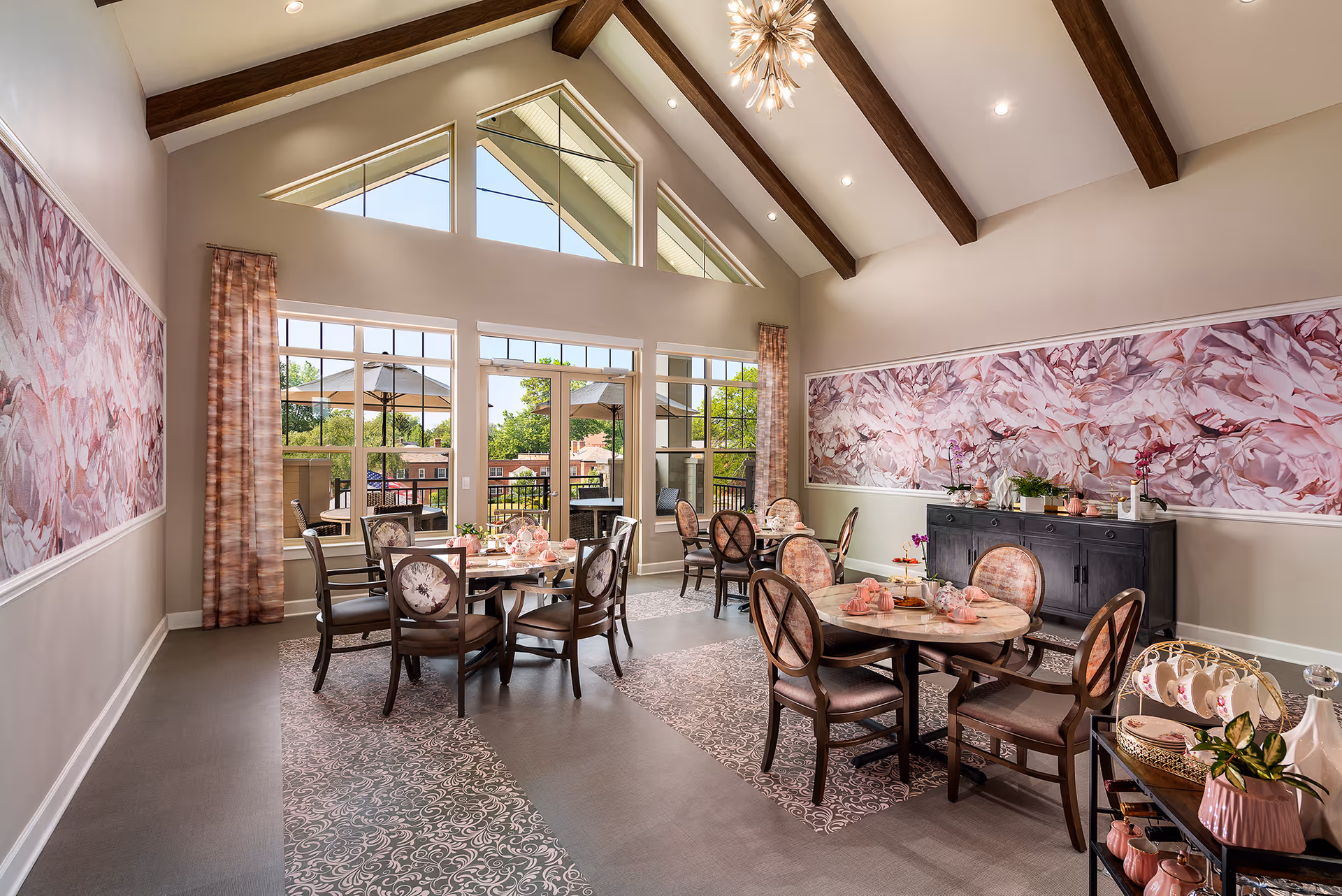 A bright dining room with high vaulted ceilings featuring exposed wooden beams and a modern chandelier. The room has large windows and glass doors leading to an outdoor patio with umbrellas. There are round tables with floral-patterned chairs, set with pink tableware and decorations. The walls are adorned with large floral artwork, and a black sideboard holds plants and decorative items.