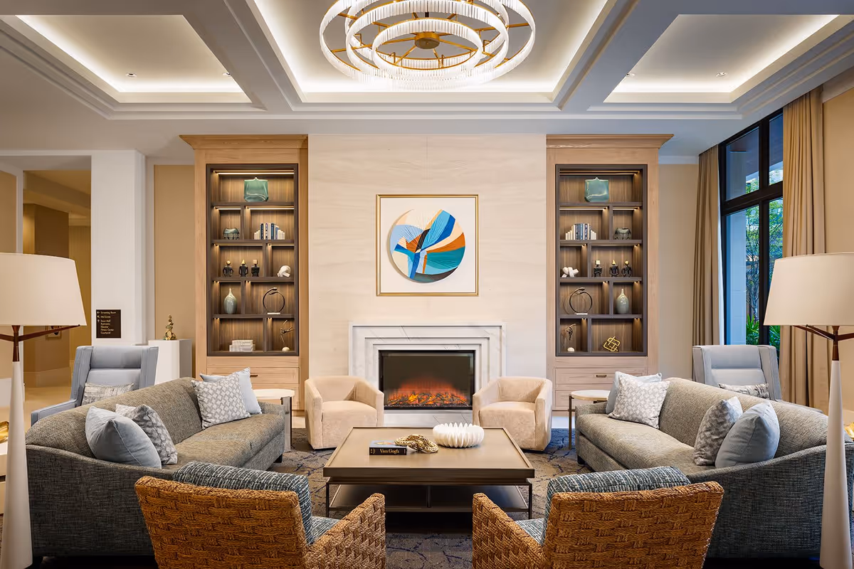 Bright, upscale lounge with sofas and armchairs arranged around a central coffee table and fireplace, flanked by built-in shelving and large windows.