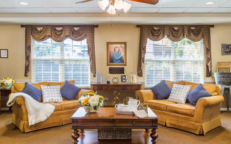 A cozy living room with two mustard yellow sofas adorned with blue and white patterned pillows. A wooden coffee table with a flower arrangement, a pitcher, cups, and a book sits in the center. Behind the sofas are two large windows with brown valances and blinds. A framed portrait of an elderly woman hangs on the wall above a wooden console table with decorative items. The room has a warm and inviting atmosphere.