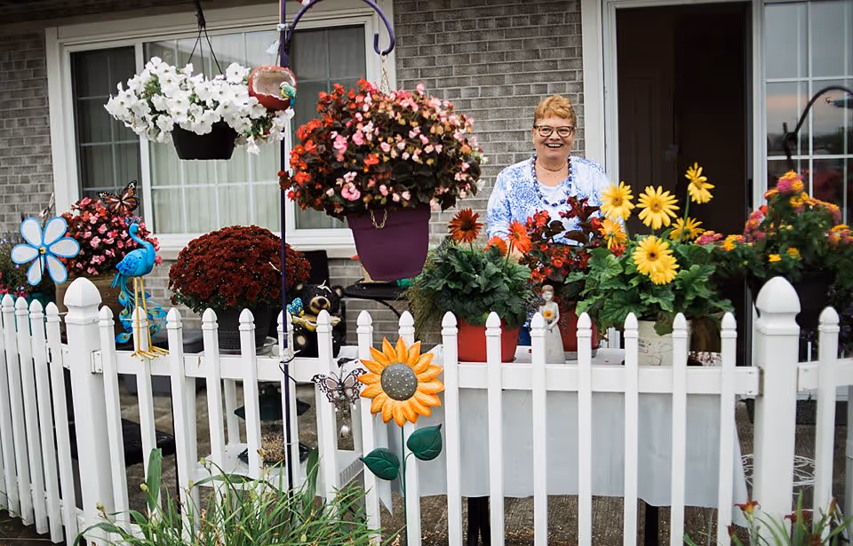A smiling woman standing behind a white picket fence adorned with colorful flowers and garden decorations in front of a brick building with windows and a door.