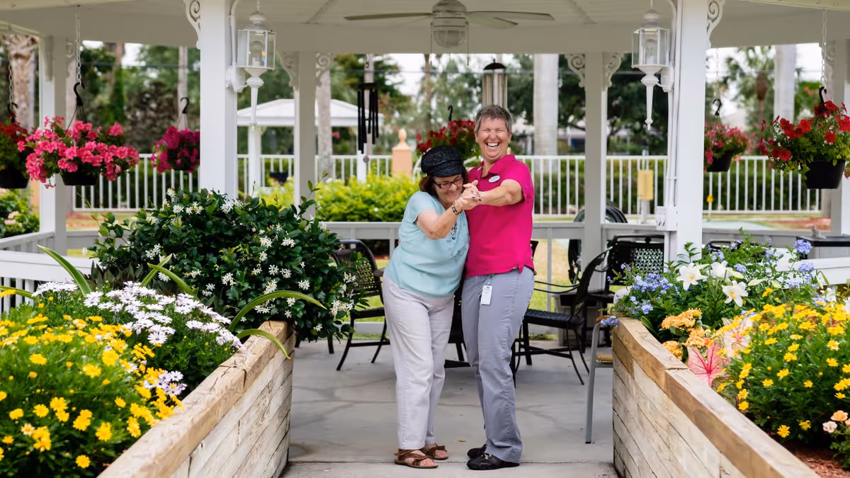 Two women smiling and holding hands while standing in a covered outdoor garden area with colorful flowers and hanging plants around them.