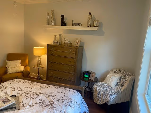 A cozy bedroom featuring a bed with a floral patterned comforter, a wooden dresser with decorative items on top, a small round side table with a lamp, and two armchairs with pillows and a throw blanket. The walls are light-colored, and there is a window letting in natural light.