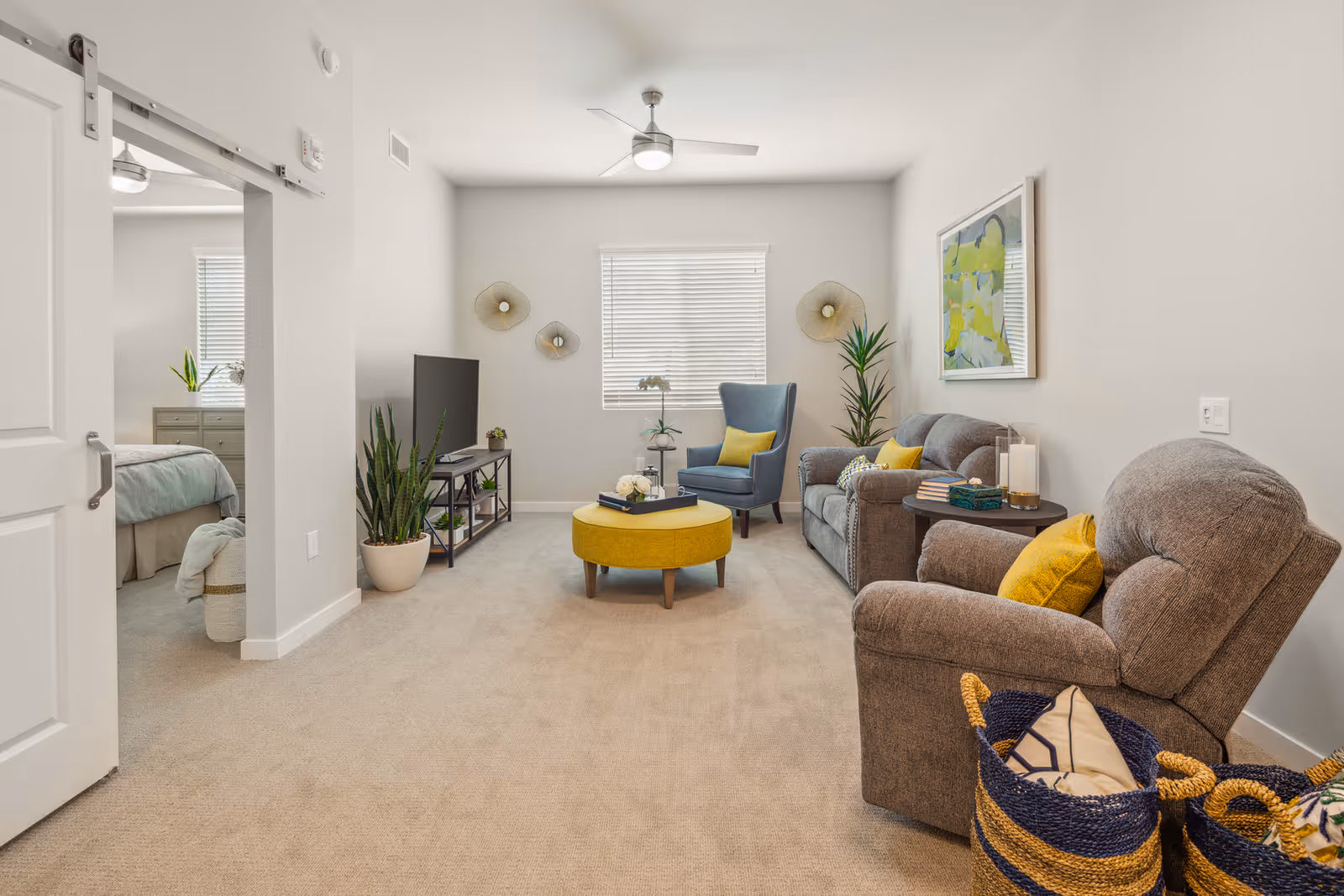 Light-filled living room with gray upholstered seating, a yellow ottoman, TV stand, and a view into an adjacent bedroom.