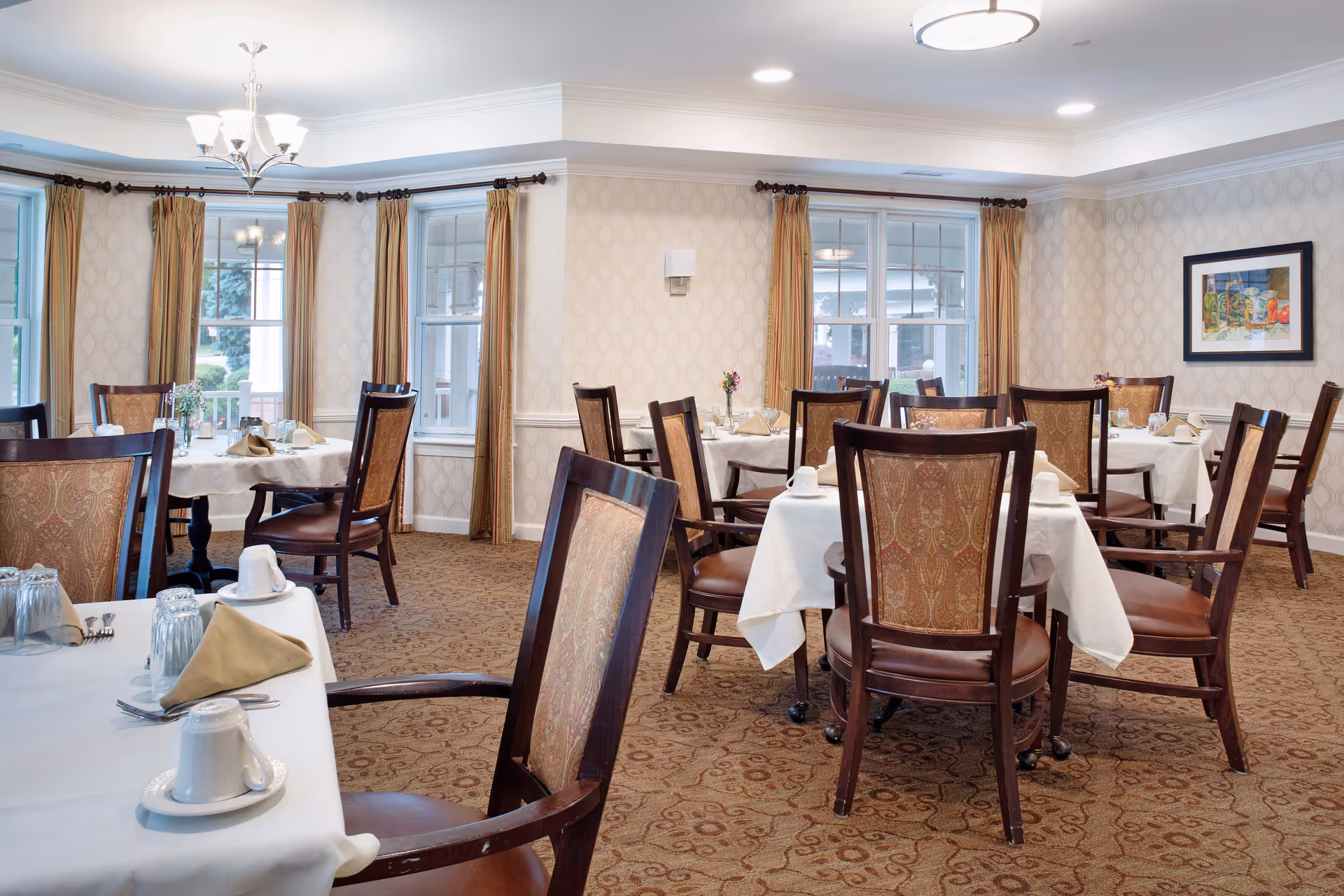 A dining room in a senior living facility with multiple round tables covered with white tablecloths, each set with cups, glasses, napkins, and silverware. The room has patterned carpet, beige wallpaper, large windows with gold curtains, and wooden chairs with upholstered backs. A chandelier and ceiling light fixtures illuminate the space.