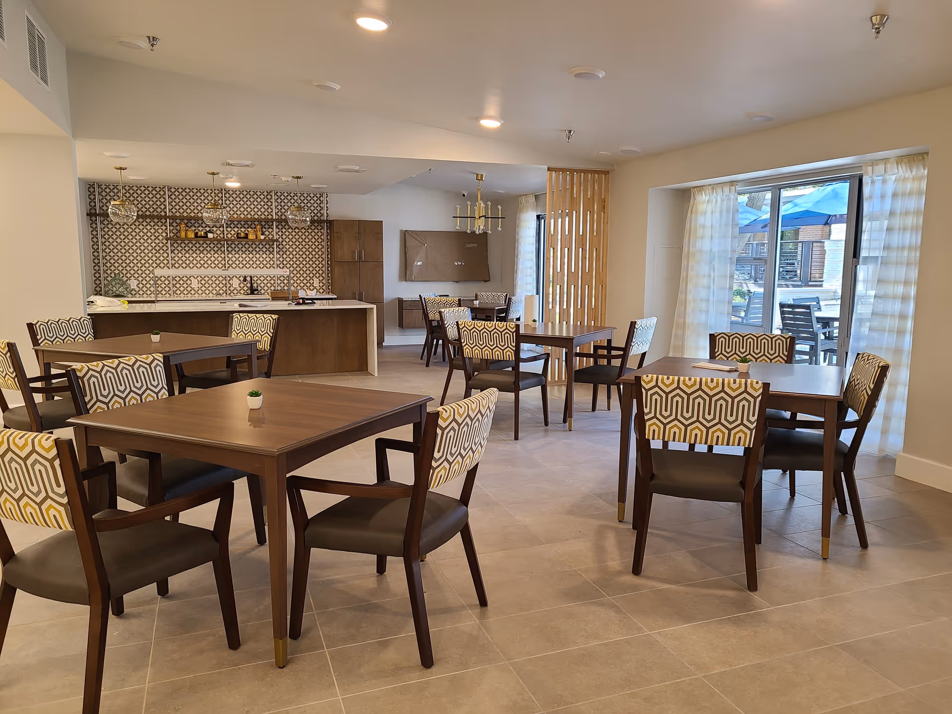 Bright communal dining area with multiple wooden tables and patterned chairs, a kitchenette/bar in the back, and large windows letting in natural light.