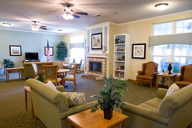 A spacious living room area in a senior living facility with multiple seating arrangements including sofas and armchairs, a fireplace with artwork above it, built-in shelves, large windows letting in natural light, ceiling fans, and an American flag in the background.