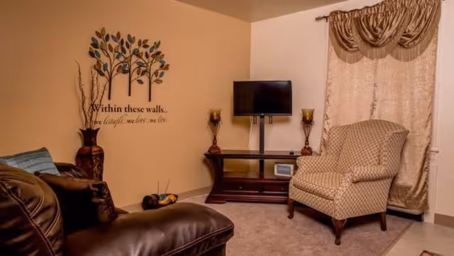 A cozy living room with a brown leather sofa on the left and a patterned armchair on the right. A flat-screen TV is mounted on a stand against the wall, flanked by two decorative candle holders. The wall has a decorative tree decal with the words 'Within these walls... we laugh, we love, we live.' Beige curtains cover a window on the right side.