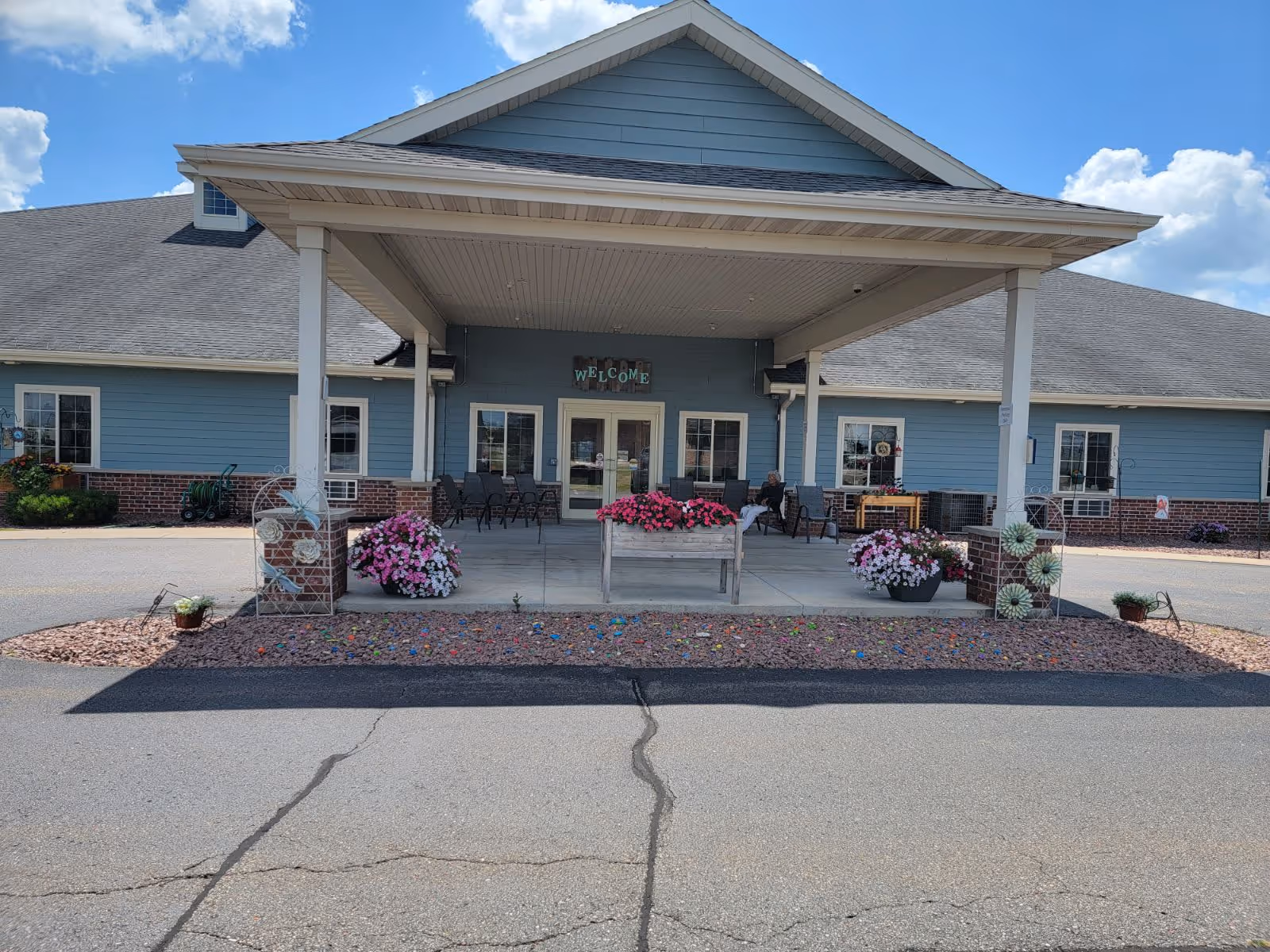 Front entrance of a single-story building with a covered porch supported by white columns. The building has blue siding with a brick base and a gray shingled roof. There are several chairs on the porch, flower pots with pink and white flowers, and a 'WELCOME' sign above the double glass doors. The sky is partly cloudy.