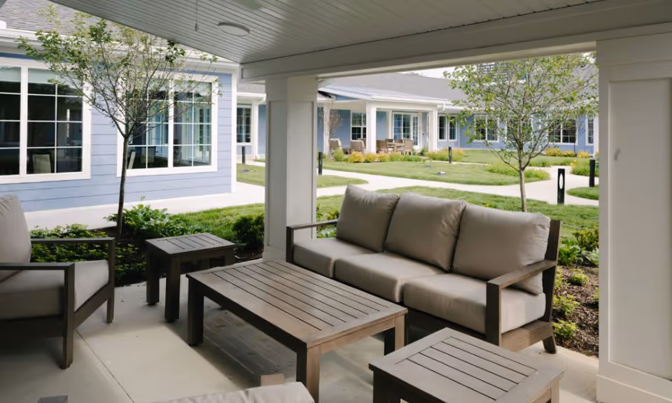 Covered outdoor seating area with cushioned chairs and a sofa around wooden tables, overlooking a landscaped courtyard with trees and pathways, surrounded by light blue buildings with white trim.