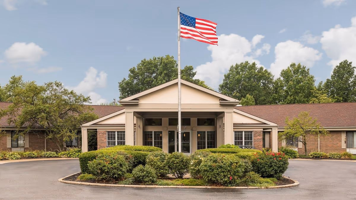 Front entrance of a single-story brick senior living building with an American flag on a flagpole and a landscaped circular driveway.
