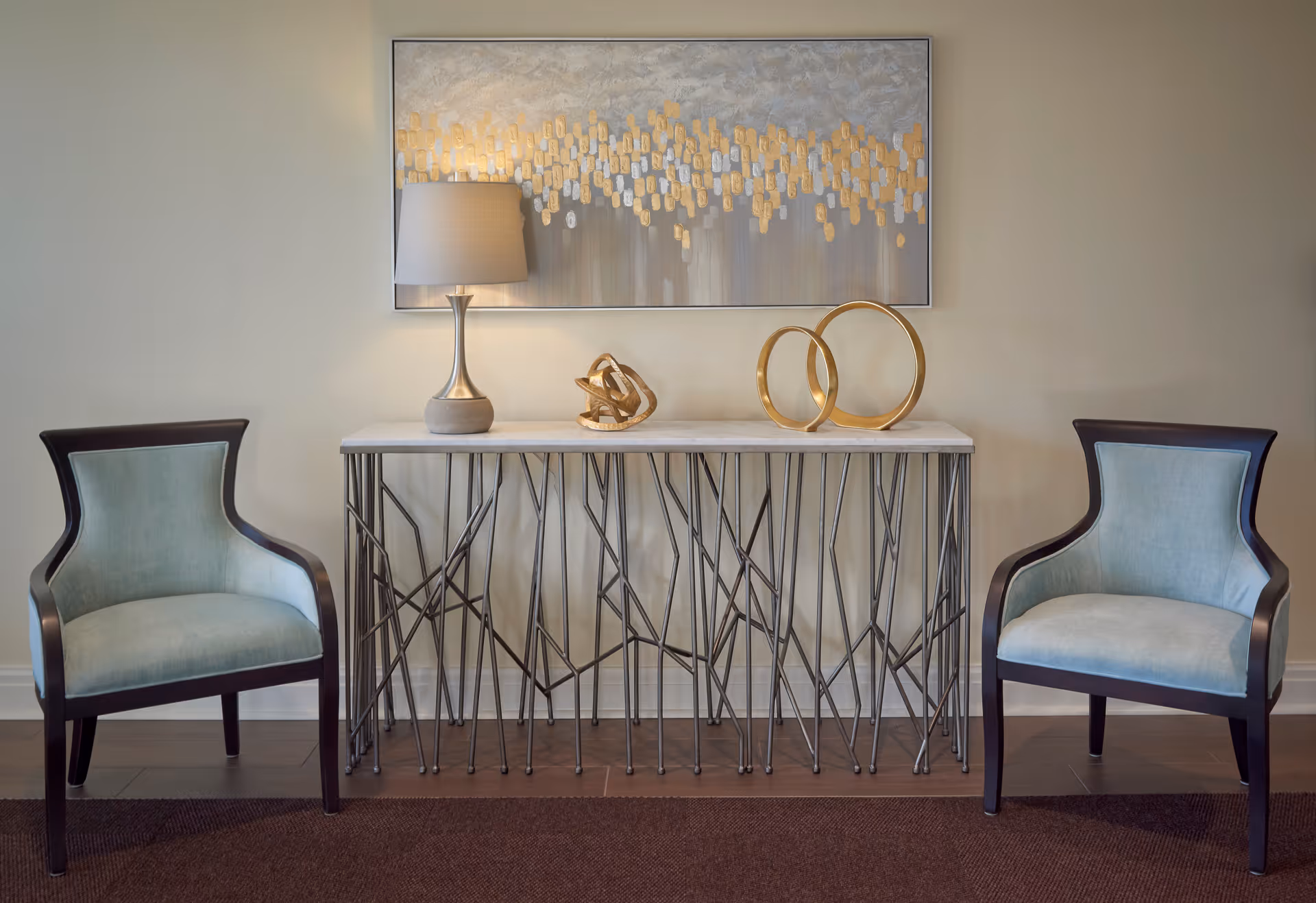 Two upholstered armchairs flank a decorative metal console table topped with a lamp, sculptures, and a painting on the wall.
