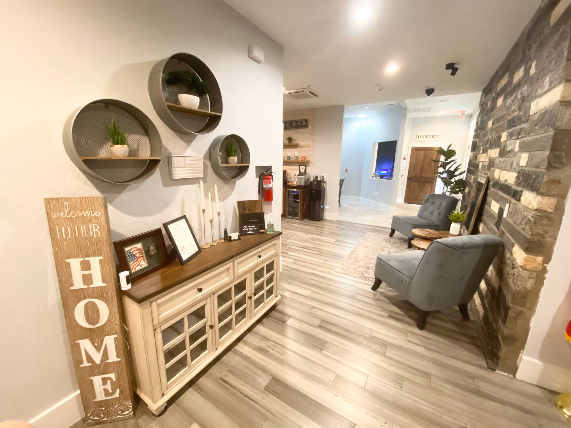 Bright interior lobby/common area with a console table and wall decor on the left, two upholstered chairs by a stone accent wall on the right, and a pantry door in the background.