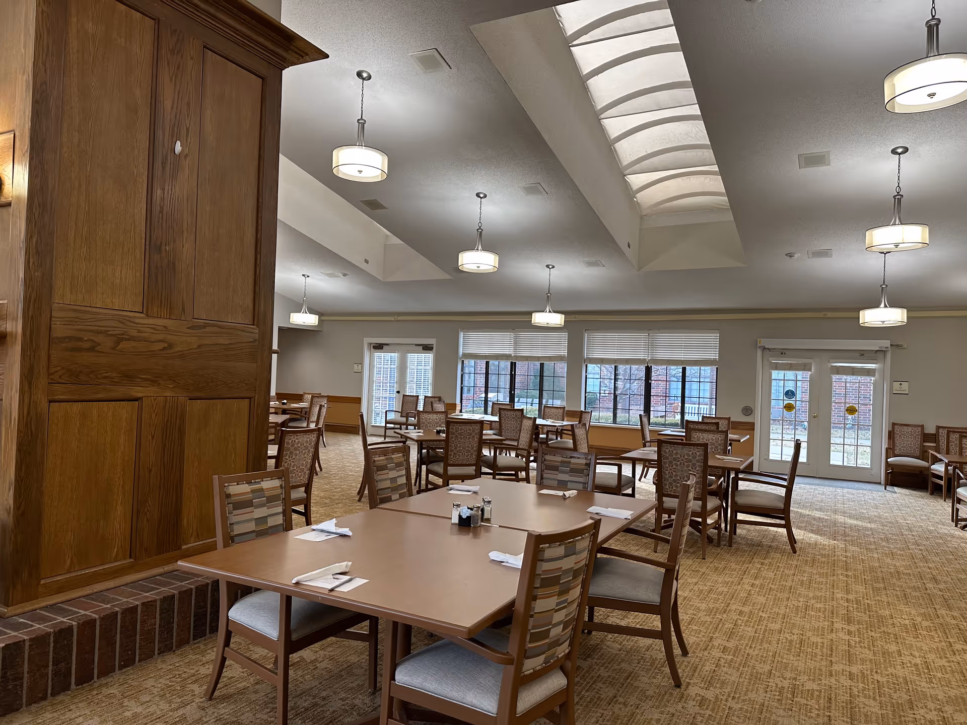 A spacious dining room with multiple wooden tables and chairs arranged neatly. The room has large windows letting in natural light, beige carpeted flooring, and several ceiling lights. There is a wooden column on the left side and double glass doors at the far end of the room.