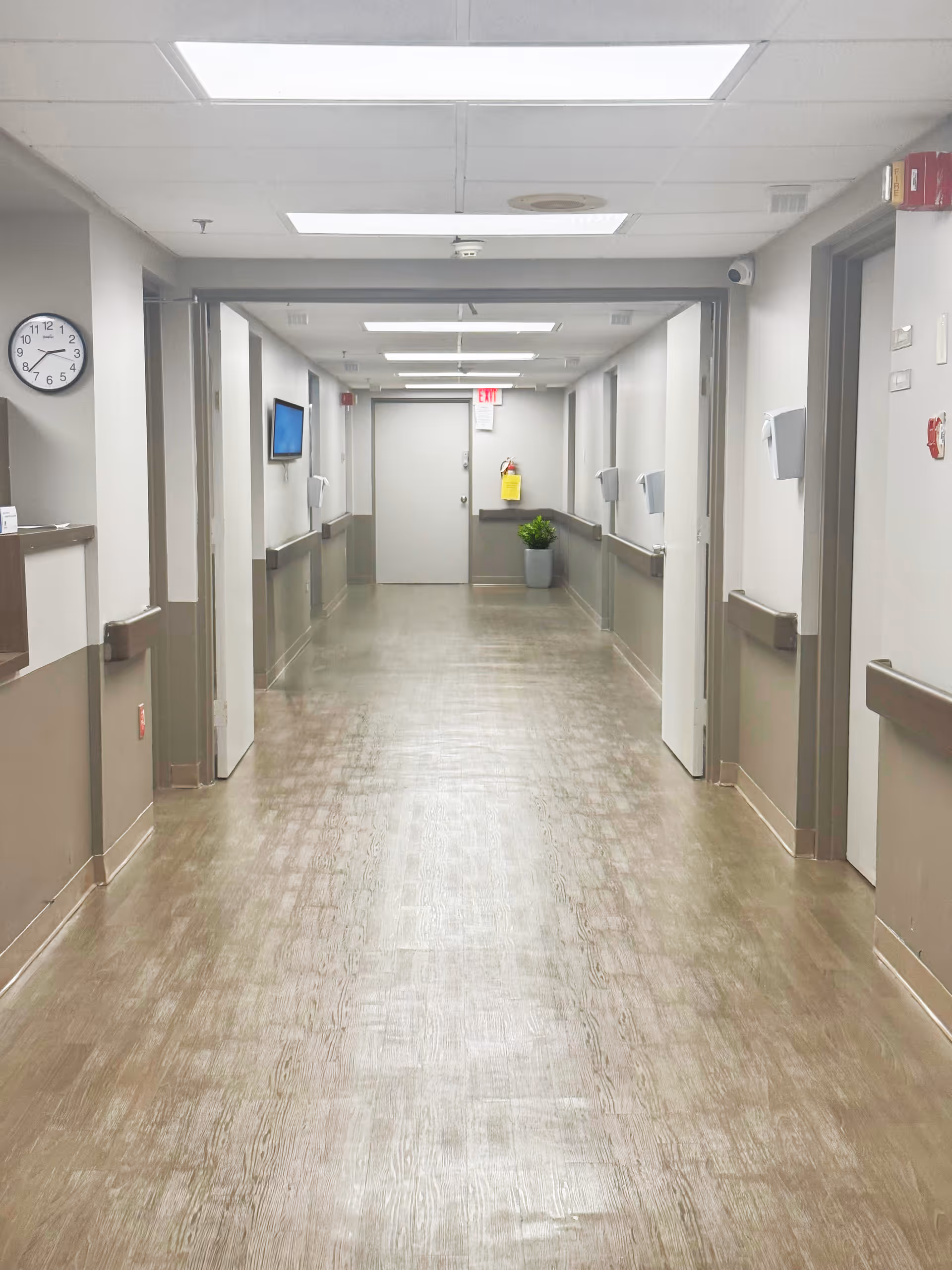 A clean, well-lit hallway in a senior living facility with beige walls and wood-patterned flooring. The hallway has multiple doors on both sides, handrails along the walls, a clock on the left wall, a small potted plant at the end near a closed door, and ceiling lights evenly spaced.