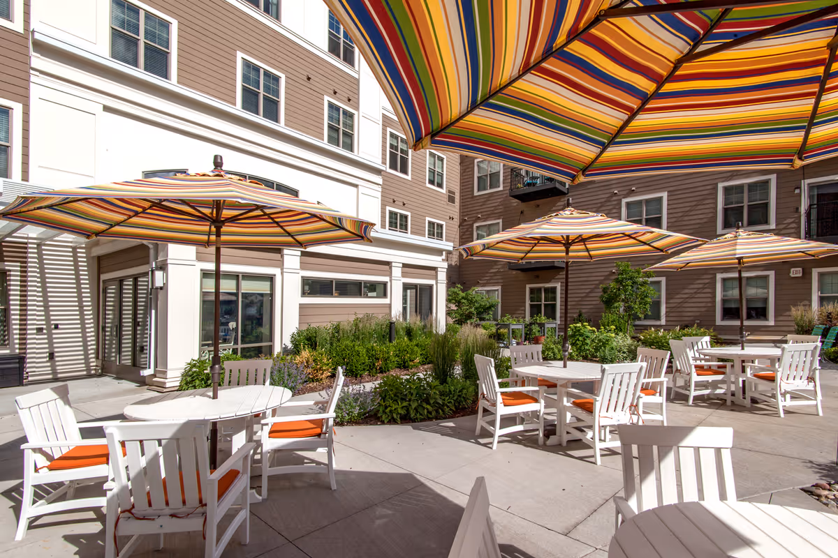Outdoor patio area with several white tables and chairs featuring orange cushions, shaded by large striped umbrellas. The patio is surrounded by a multi-story residential building with windows and some greenery along the edges.