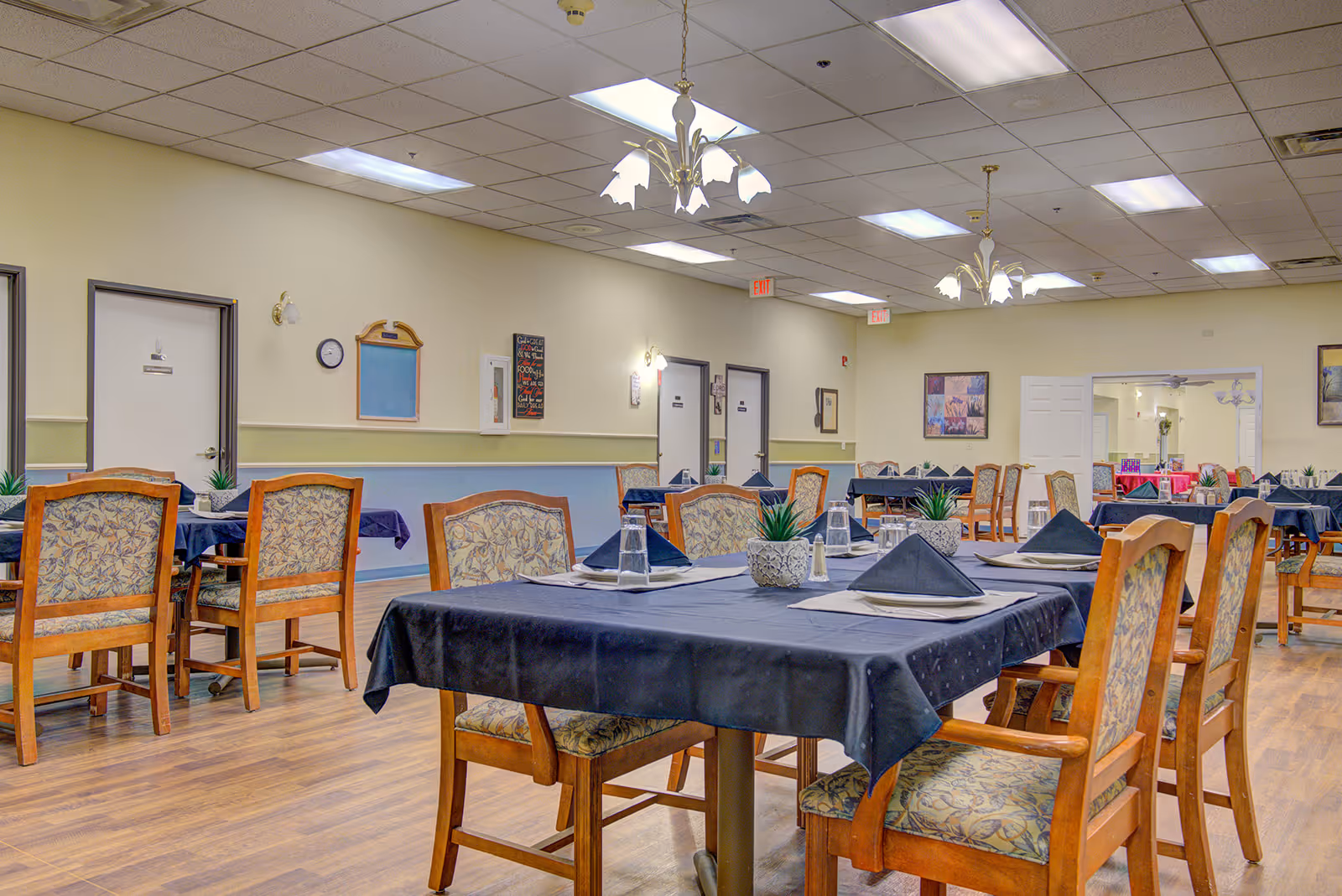 Spacious senior dining room with multiple tables set with black tablecloths, wooden chairs, and overhead chandeliers.