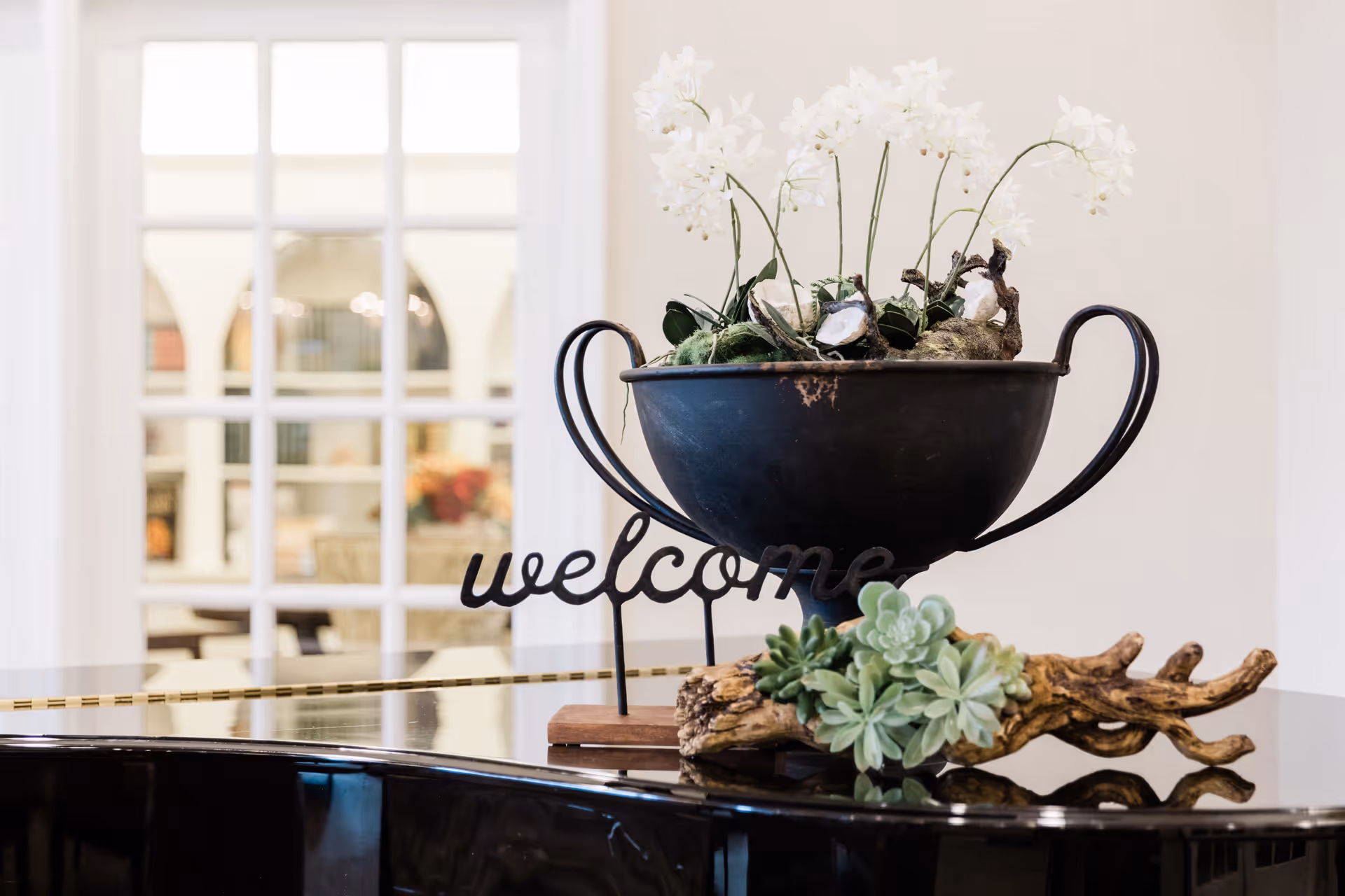 A decorative arrangement on a glossy black surface featuring a black metal bowl with white flowers and a wooden piece with green succulents, accompanied by a metal sign that says 'welcome'. In the background, there is a white-framed glass door leading to a room with shelves and furniture.