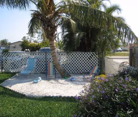 Sand play area with palm tree, colorful chairs, and lattice fence in a sunny yard.