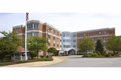 Exterior view of a multi-story healthcare facility building with a curved driveway, landscaped greenery, and an American flag on a flagpole near the entrance.