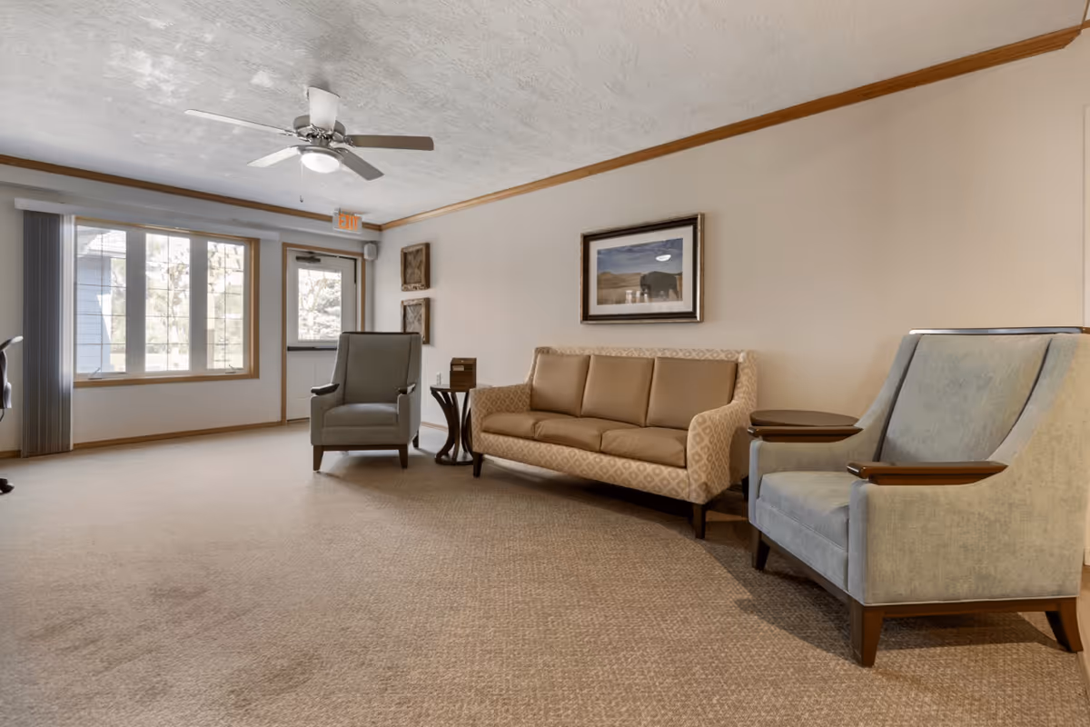 A senior living facility common area with beige carpet, a beige patterned sofa, two blue armchairs with wooden armrests, a small round side table, a ceiling fan, and a large window letting in natural light. There is a framed picture on the wall and an exit door near the window.