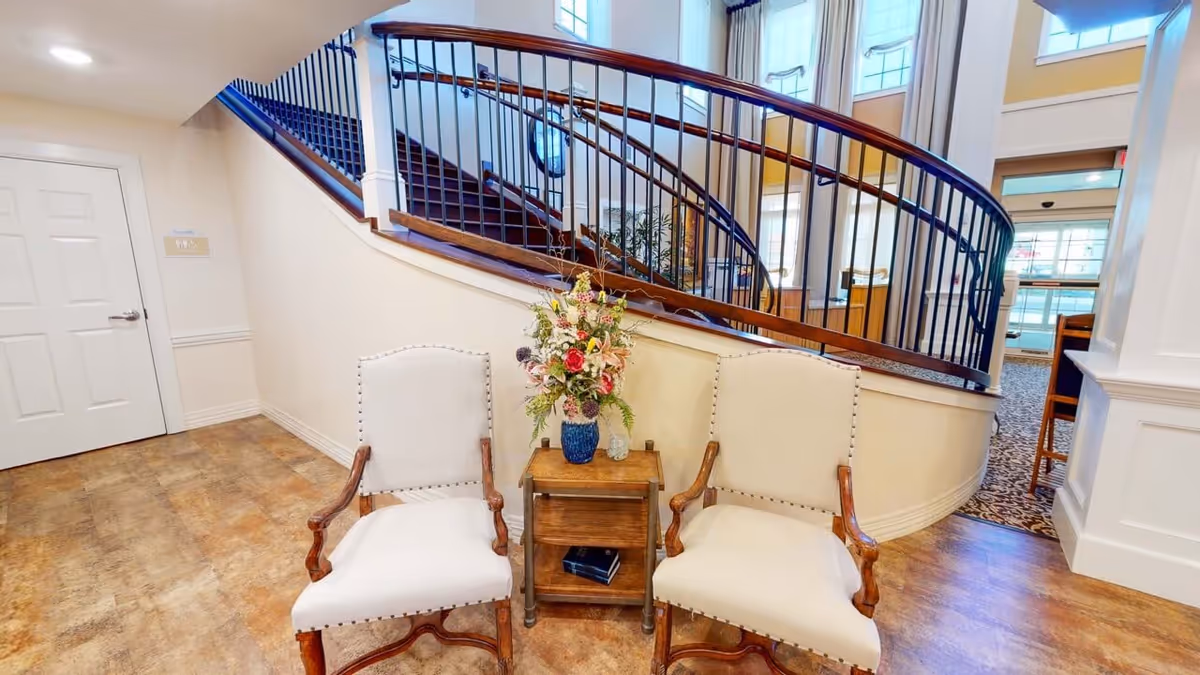 Two upholstered armchairs flank a small wooden side table with a flower arrangement in front of a curved staircase and large windows in a bright lobby.